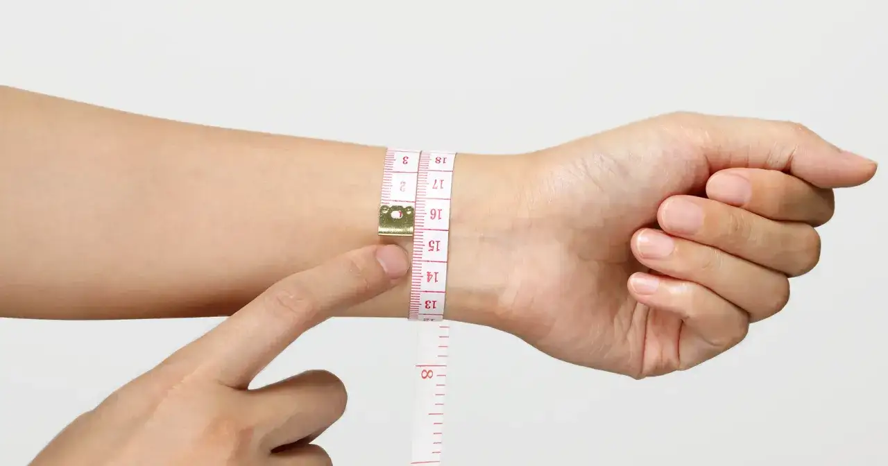 A hand uses a measuring tape to determine wrist size, showing numbers like 13, 14, 15, 16, 17, 18, useful for a bracelet size chart.